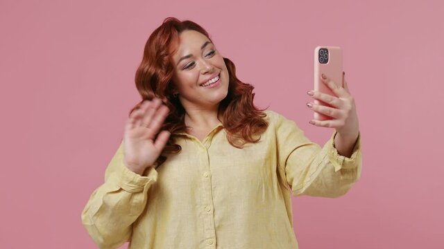Young ginger overweight woman 20s wears yellow shirt get video call using mobile cell phone do selfie talk conduct pleasant conversation greet with hand isolated plain pink background studio portrait
