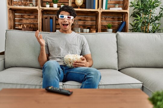 Young hispanic man watching movie using 3d glasses sitting on the sofa at home.