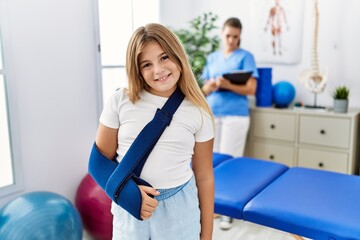 Blonde little girl wearing arm on sling at rehabilitation clinic looking positive and happy...