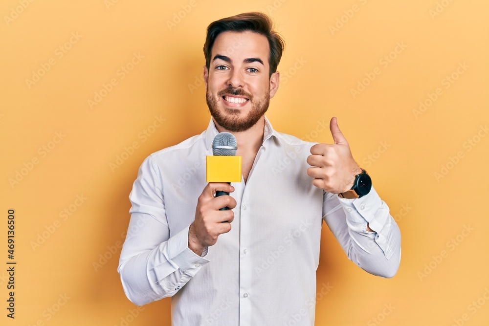 Wall mural Handsome caucasian man with beard holding reporter microphone smiling happy and positive, thumb up doing excellent and approval sign