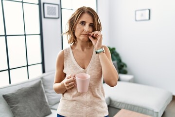 Middle age woman drinking a cup coffee at home mouth and lips shut as zip with fingers. secret and...