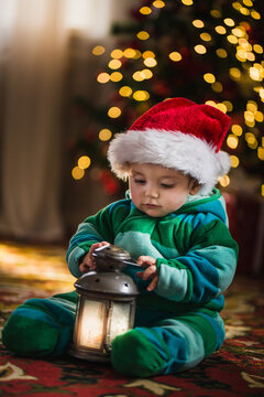 Little baby boy in Santa Claus hat sits under Christmas tree and plays vintage lantern