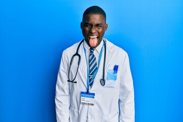 Young african american man wearing doctor uniform sticking tongue out happy with funny expression. emotion concept.
