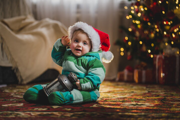 Little baby boy in Santa Claus hat sits under Christmas tree and plays vintage lantern