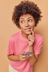 Image of curly haired woman bites lips has thoughtful expression looks away types message content dressed in casual t shirt isolated over beige background checks social media has gadget addiction