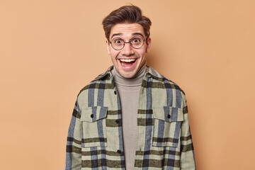 Fototapeta premium Indoor shot of happy handsome European man looks with cheerful surprised expression hears great news enjoys good day wears round spectacles and checkered shirt isolated over beige studio wall