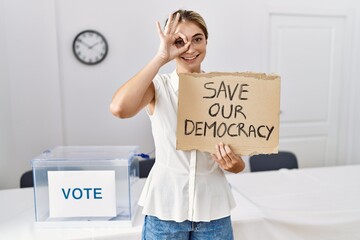 Young blonde woman at political election holding save out democracy banner smiling happy doing ok sign with hand on eye looking through fingers