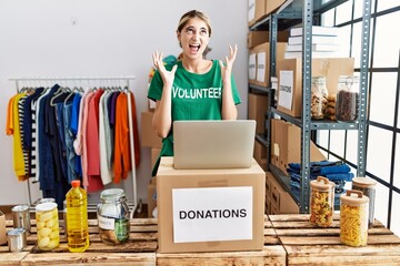 Young blonde woman wearing volunteer t shirt at donations stand crazy and mad shouting and yelling with aggressive expression and arms raised. frustration concept.