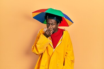 Young african american man wearing yellow raincoat smelling something stinky and disgusting, intolerable smell, holding breath with fingers on nose. bad smell