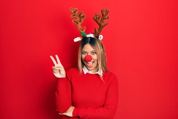 Beautiful hispanic woman wearing deer christmas hat and red nose smiling with happy face winking at...