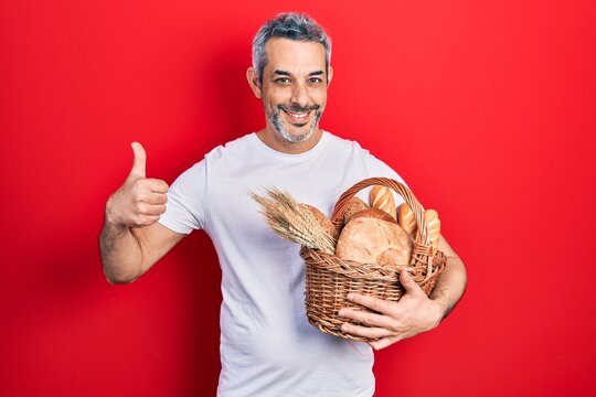 Handsome middle age man with grey hair holding wicker basket with bread smiling happy and positive, thumb up doing excellent and approval sign