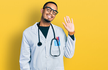 Young african american man wearing doctor uniform and stethoscope waiving saying hello happy and smiling, friendly welcome gesture