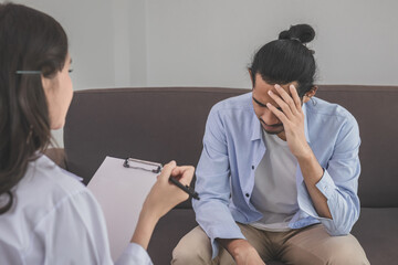 Asian young man, male has mental symptoms which must have been therapy and stress, sitting on couch to consult to psychologist during the session  taking notes to find out how to treat the therapist.