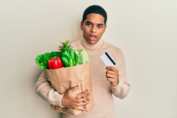 Young handsome hispanic man holding groceries and credit card in shock face, looking skeptical and sarcastic, surprised with open mouth