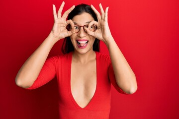 Young latin woman wearing casual clothes and glasses doing ok gesture like binoculars sticking tongue out, eyes looking through fingers. crazy expression.