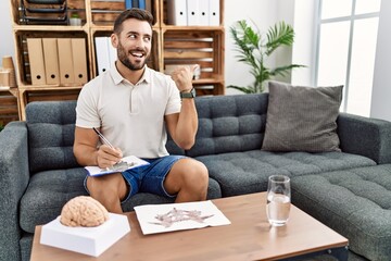 Handsome hispanic man working with rorschach test at psychology clinic smiling with happy face looking and pointing to the side with thumb up.