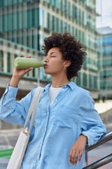 Vertical shot of curly haired woman feels thirsty drinks fresh water from bottle keeps eyes closed...