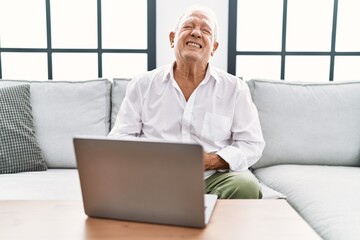 Senior man using laptop at home sitting on the sofa with hand on stomach because indigestion,...
