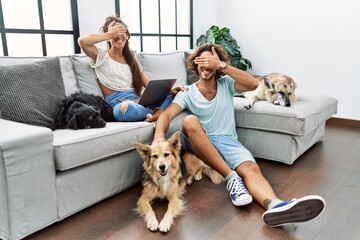 Young hispanic couple with dogs relaxing at home smiling and laughing with hand on face covering eyes for surprise. blind concept.