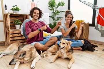Young hispanic couple doing laundry with dogs smiling and looking at the camera pointing with two hands and fingers to the side.