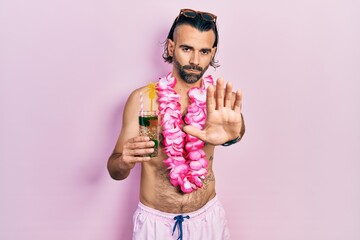 Young hispanic man wearing swimsuit and hawaiian lei drinking tropical cocktail doing stop sing with palm of the hand. warning expression with negative and serious gesture on the face.