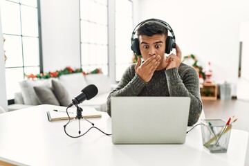 Handsome hispanic man recording podcast covering mouth with hand, shocked and afraid for mistake....