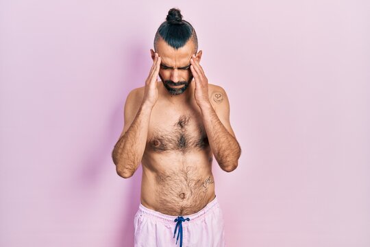 Young hispanic man shirtless wearing swimsuit with hand on head for pain in head because stress. suffering migraine.