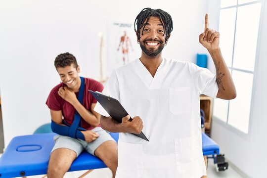 Young hispanic man working at pain recovery clinic with a man with broken arm smiling amazed and surprised and pointing up with fingers and raised arms.