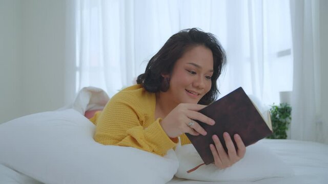 Asian woman oversize big chubby girl lying in bed. She wearing a yellow shirt reads books, novels, fairy tales, smiles and laughs happily. hobbies on holiday.
