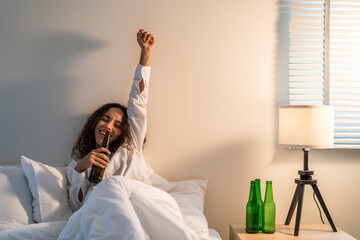 Drunk Latino woman hold beer bottle, feel hangover and dancing on bed. - obrazy, fototapety, plakaty