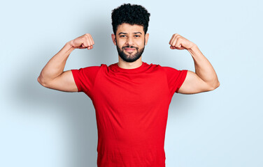 Young arab man with beard wearing casual red t shirt showing arms muscles smiling proud. fitness concept.