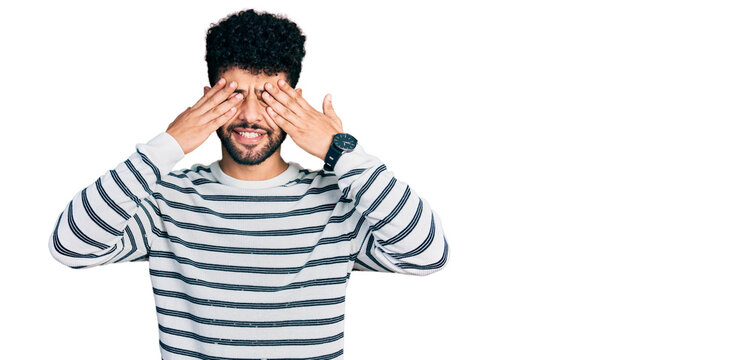 Young arab man with beard wearing casual striped sweater covering eyes with hands smiling cheerful and funny. blind concept.