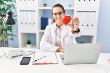 Young hispanic woman wearing doctor uniform holding heart at clinic