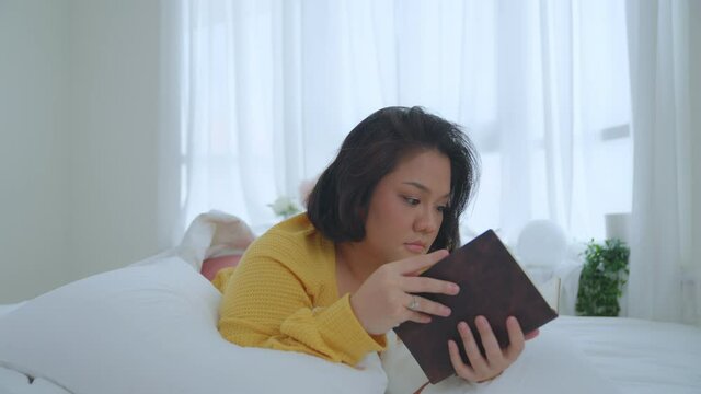 Asian woman oversize big chubby girl lying in bed reading a book in the bedroom showing a panic look at the camera.