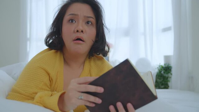 Asian woman oversize big chubby girl lying in bed reading a book in the bedroom showing a panic look at the camera. Leisure and hobbies on vacation. 
