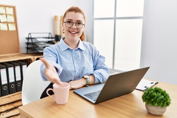 Young redhead woman working at the office using computer laptop smiling friendly offering handshake as greeting and welcoming. successful business.