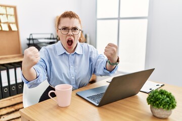Young redhead woman working at the office using computer laptop angry and mad raising fists frustrated and furious while shouting with anger. rage and aggressive concept.
