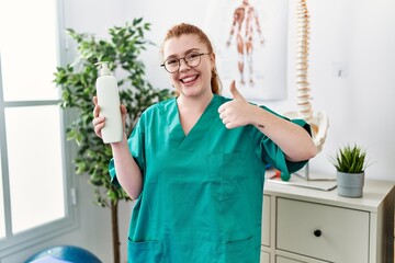 Young redhead physiotherapist woman holding massage body lotion smiling happy and positive, thumb up doing excellent and approval sign