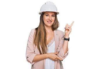 Young blonde woman wearing architect hardhat smiling happy pointing with hand and finger to the side