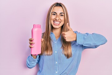 Beautiful hispanic woman holding shampoo bottle smiling happy and positive, thumb up doing excellent and approval sign