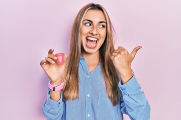 Beautiful hispanic woman holding menstrual cup pointing thumb up to the side smiling happy with open mouth