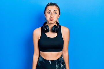 Beautiful hispanic woman wearing gym clothes and using headphones puffing cheeks with funny face....