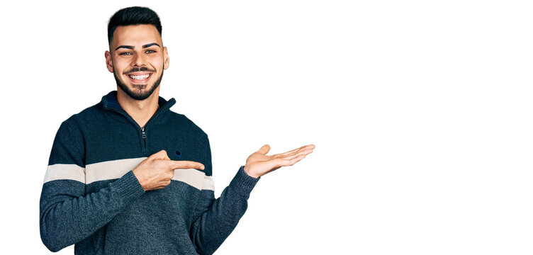 Young hispanic man with beard wearing casual winter sweater amazed and smiling to the camera while presenting with hand and pointing with finger.