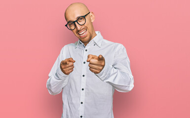 Bald man with beard wearing business shirt and glasses pointing fingers to camera with happy and funny face. good energy and vibes.