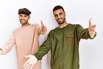 Young gay couple standing over isolated white background looking at the camera smiling with open arms for hug. cheerful expression embracing happiness.