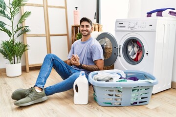 Young hispanic man putting dirty laundry into washing machine with hands together and crossed fingers smiling relaxed and cheerful. success and optimistic