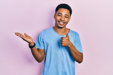 Fototapeta premium Young african american man wearing casual blue t shirt showing palm hand and doing ok gesture with thumbs up, smiling happy and cheerful