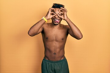 Young african american man wearing swimwear and swimmer glasses doing ok gesture like binoculars sticking tongue out, eyes looking through fingers. crazy expression.