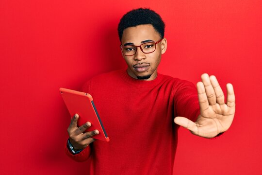 Young african american man using touchpad device with open hand doing stop sign with serious and confident expression, defense gesture