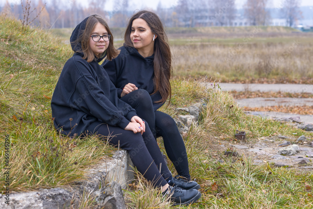 Wall mural Two beautiful girls with long hair in black clothes sit together on the grass on the outskirts of the city.